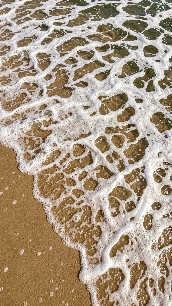 Overhead view of lacy white ocean foam spreading across wet tan beach sand