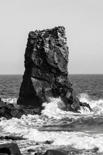 Black and white view of a tall sea stack with waves crashing against its rocky base