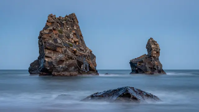 Long exposure shot of jagged sea stacks rising from smooth calm ocean under a pale blue sky at blue hour