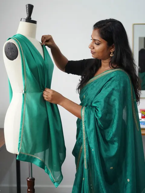 A South Asian seamstress adjusts emerald silk fabric on a dress form in a bright design studio.