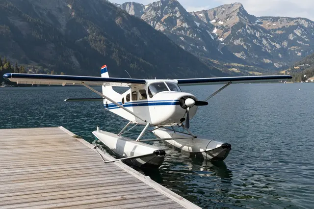 A white and blue seaplane is moored to a wooden dock on a calm alpine lake backed by rocky mountains.