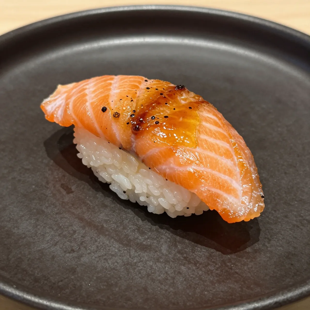 A single piece of seared caramelized salmon nigiri rests on a matte dark gray plate.
