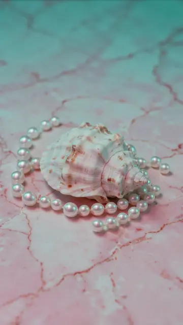A spiral seashell rests surrounded by a pearl strand on pink veined marble under soft teal lighting