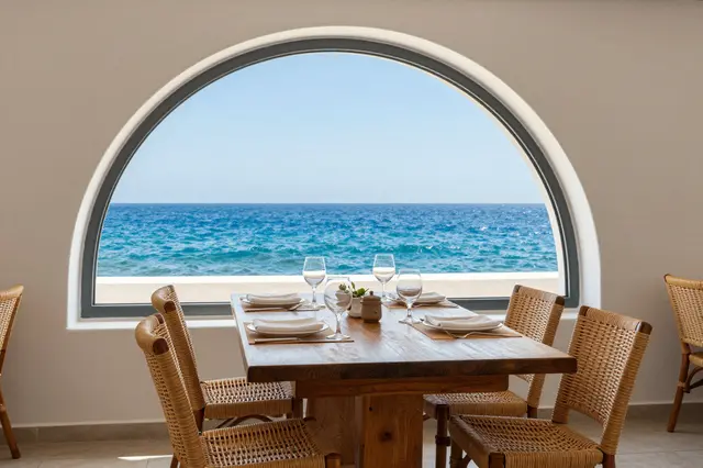 Set olive wood dining table with wicker chairs sits in front of an arched window overlooking the blue Mediterranean sea.