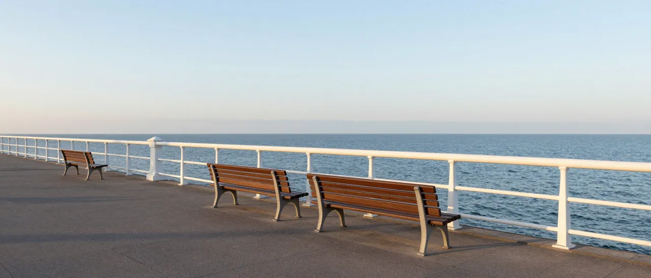 Empty wooden benches line a white railed seaside promenade overlooking calm ocean at sunrise