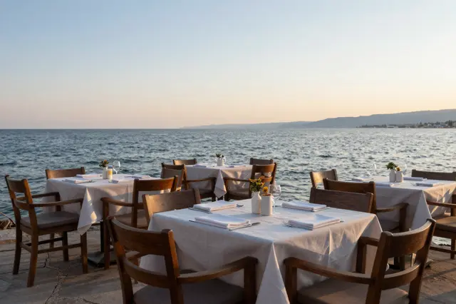 Set dining tables with white linens line a stone waterfront beside calm sea at golden sunset