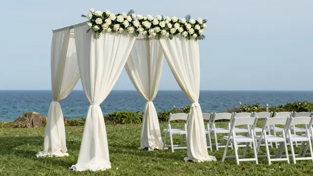 A white rose and chiffon wedding chuppah stands on a coastal lawn with rows of folding chairs facing the ocean.