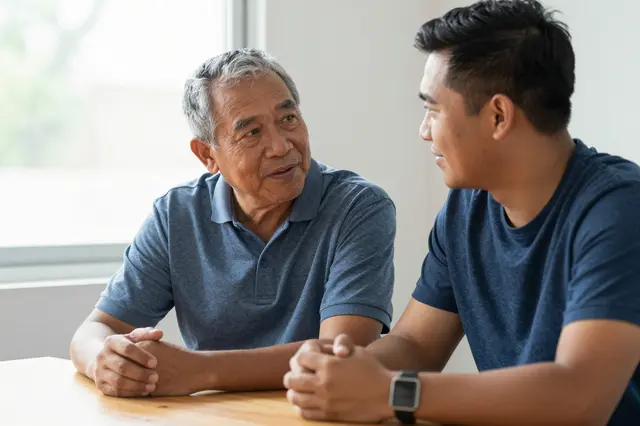 An older mixed-race man and a younger Pacific Islander man converse while seated at a sunny indoor table.