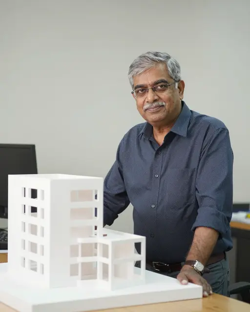South Asian senior architect leans on a table beside a white architectural building model in a bright studio
