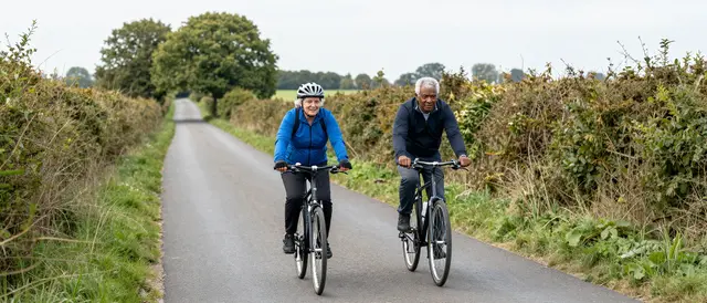 Two older adults ride bicycles side by side down a narrow hedgerow-lined rural road