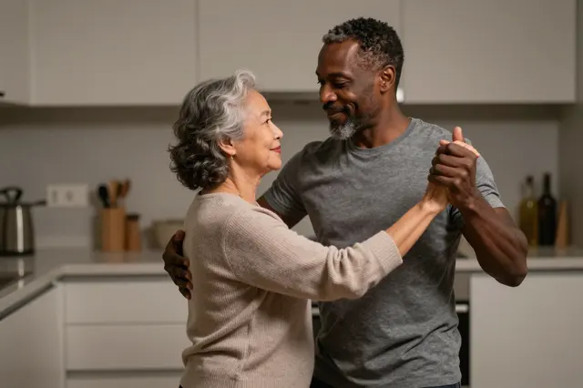 An older interracial couple slow dance together in a modern home kitchen at night, smiling gently at one another.