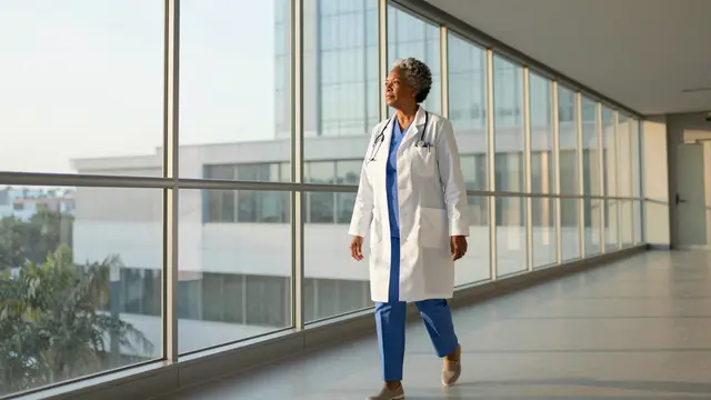 Older Black woman doctor walks along a glass hospital walkway during soft sunrise light
