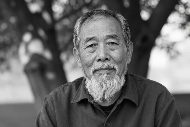 Black and white close-up portrait of an elderly East Asian man with a wispy white beard seated under a tree in dappled light