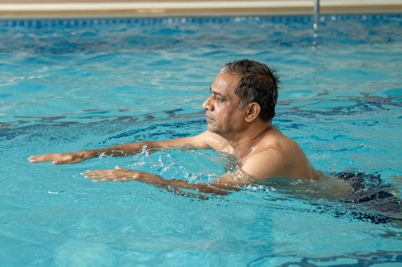 Side view of a slim older South Asian man doing water exercises in an indoor swimming pool