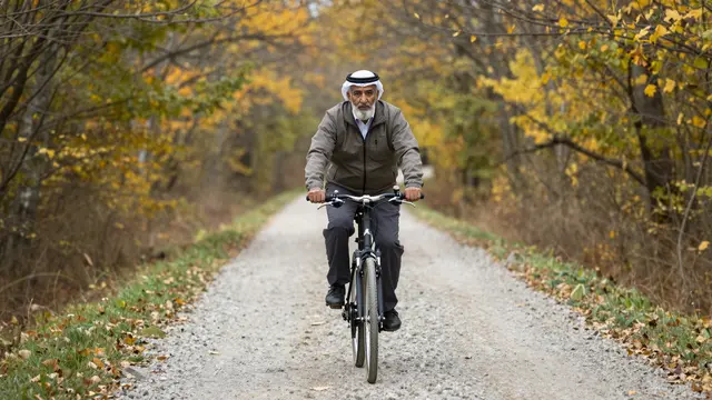 Older Middle Eastern man rides a bicycle along a gravel path through autumn woodland