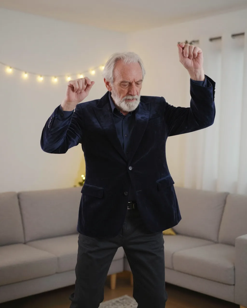 Older man in a dark velvet blazer dances in a living room lit by warm string lights