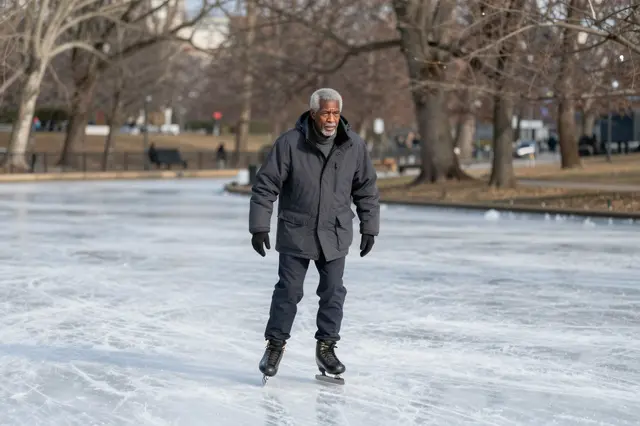 An older Black man in a winter parka ice skates on a frozen park pond lined with bare winter trees.