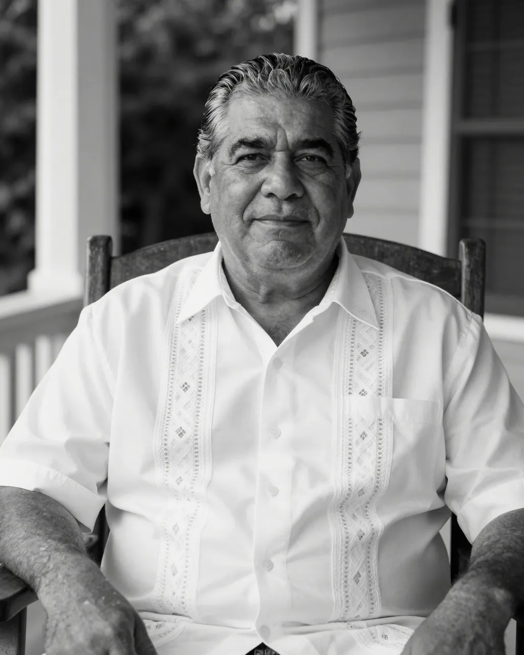 Black and white portrait of an older Latino man in a white guayabera sitting in a wooden porch chair