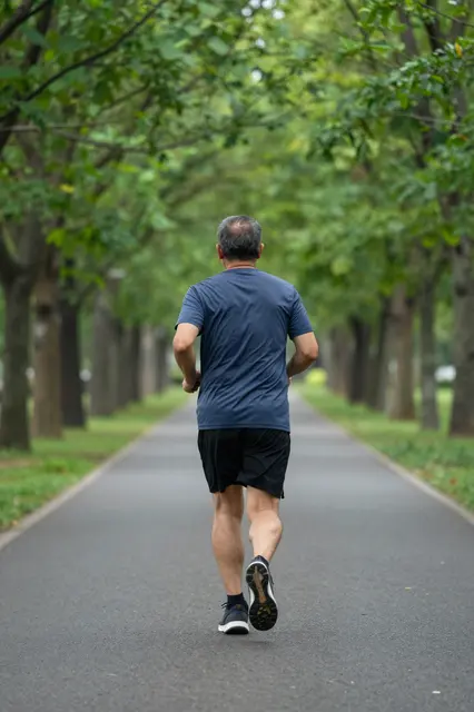 Back view of a senior Southeast Asian man jogging down a tree lined paved park path