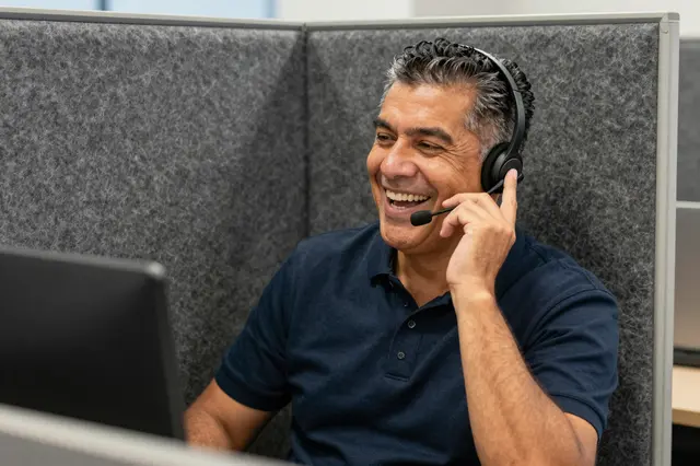 Smiling sixtysomething Latino man wears a headset while taking a call in a felt lined office booth