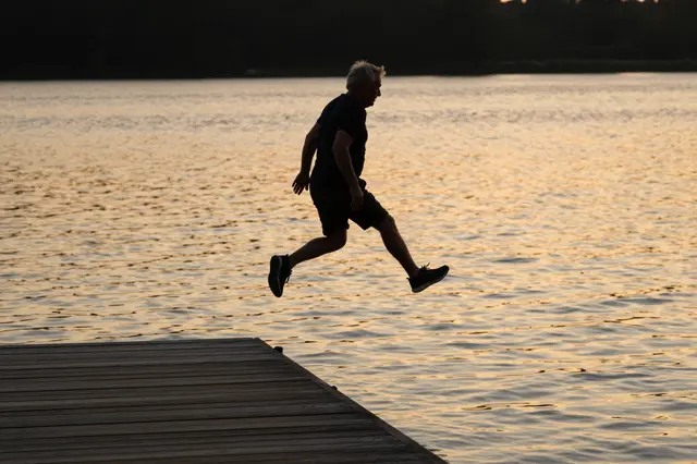Silhouette of an older man mid-leap from a wooden jetty over golden rippled lake water at sunset