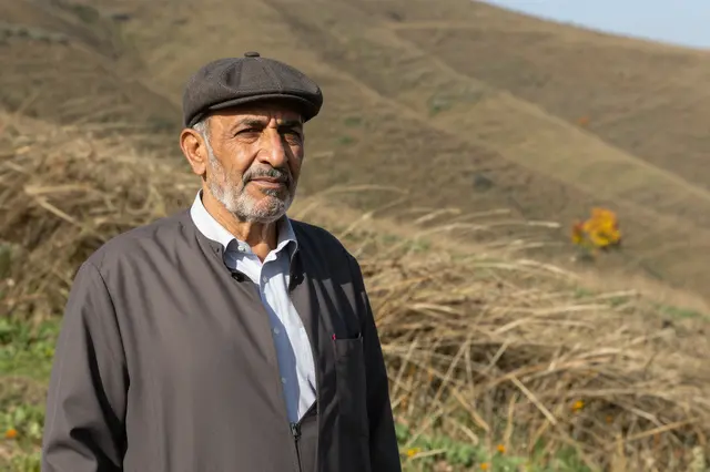Older Middle Eastern man in a flat cap stands left of frame on a dry grassy hillside
