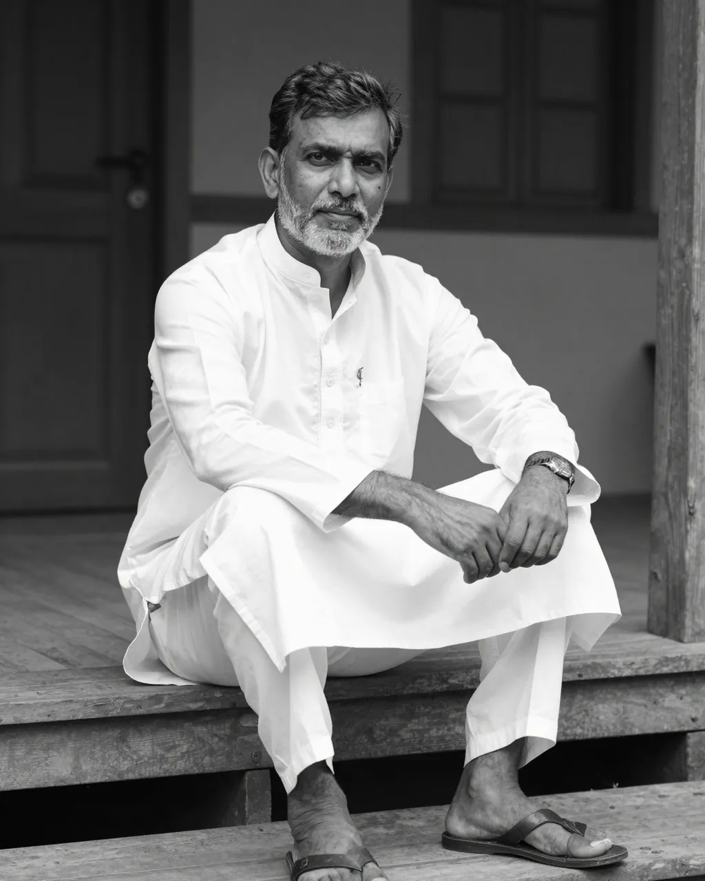 Black and white portrait of a senior South Asian man seated on wooden porch steps in a white kurta