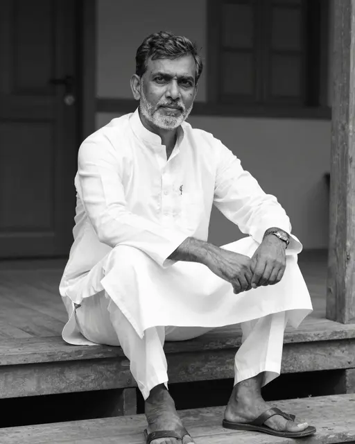 Black and white portrait of a senior South Asian man seated on wooden porch steps in a white kurta
