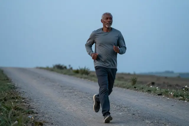 A mixed-race man in his sixties runs alone on a gravel country road in cool pre-dawn blue light.