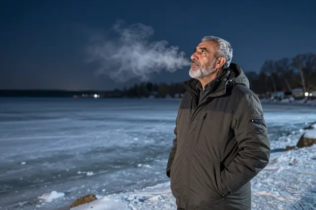 Older Middle Eastern man in a winter jacket stands on a frozen lakeshore at night with visible cold breath, looking upward