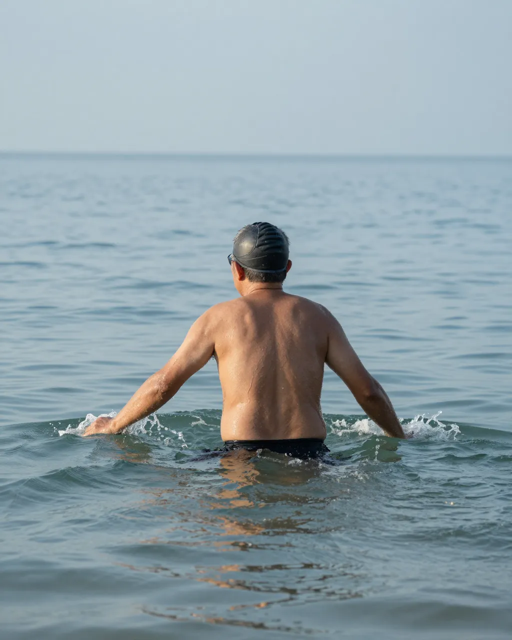 Seen from behind, an older man wades into calm pale blue sea for an early morning swim.