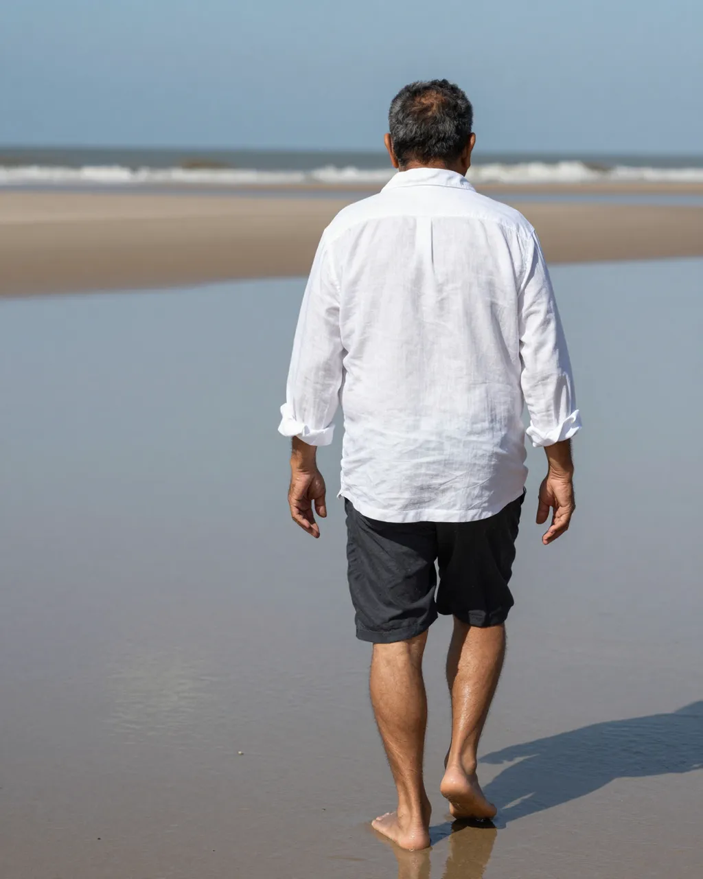 Back view of an older man in a white linen shirt walking barefoot on wet low-tide beach sand