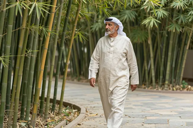 Elderly Middle Eastern man in traditional dress walks along a stone path through a bamboo grove