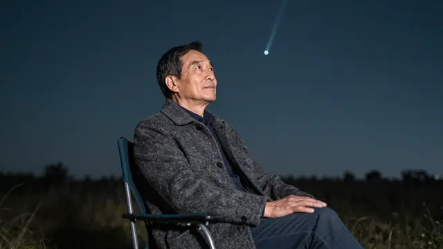 A senior East Asian man sits in a camp chair in a dark field looking up at a meteor streak in the night sky