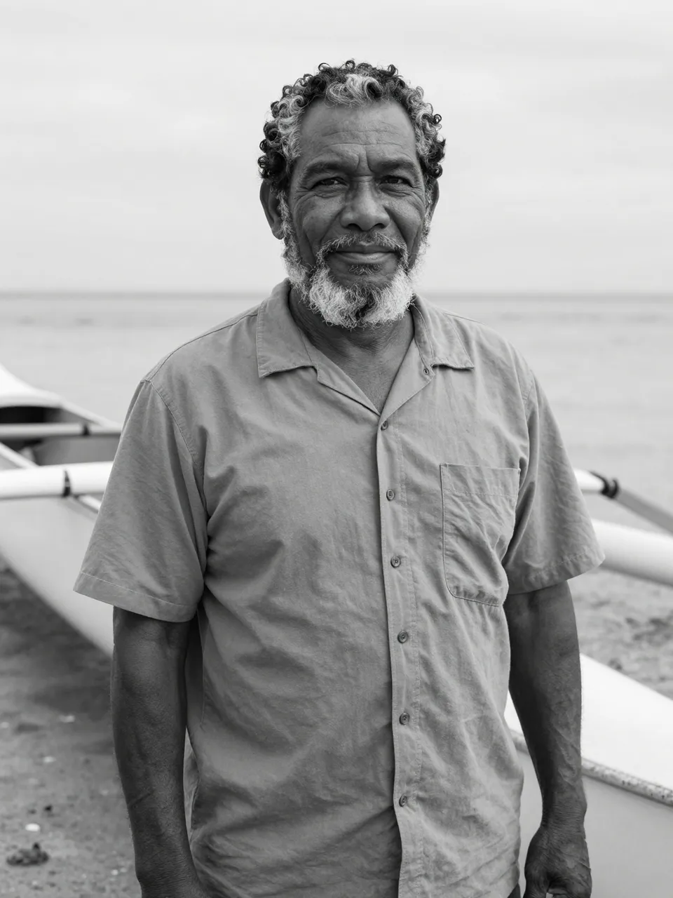 Black and white portrait of a senior bearded man standing next to an outrigger canoe on a sandy beach