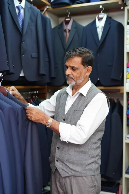 Older South Asian tailor examines hanging suits inside his small shop.