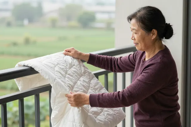 An East Asian senior woman arranges a white quilt over a balcony railing on a sunny morning
