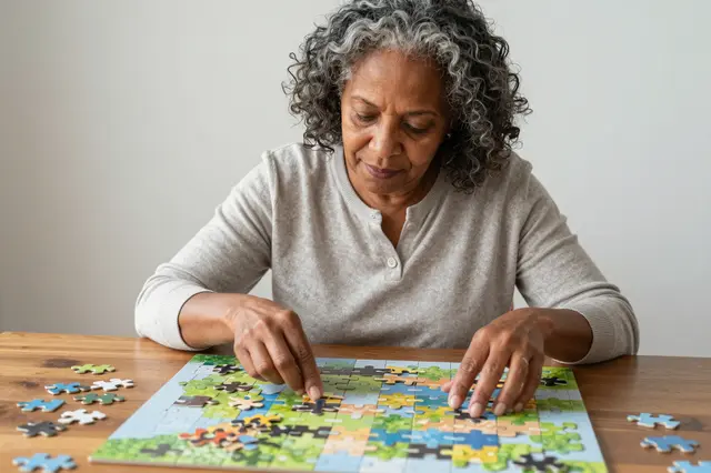 A mixed-race woman in her sixties works on a jigsaw puzzle spread across a wooden dining table.