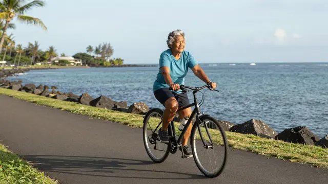 Elderly Pacific Islander woman rides a bicycle along a paved seaside promenade on a calm sunny morning.
