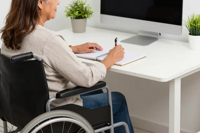 A mature Latina woman in a wheelchair writes in a notebook at a modern office desk with potted plants and a computer monitor