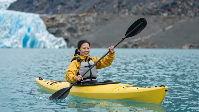 An East Asian senior woman in a yellow jacket paddles a yellow kayak on turquoise glacial lake water near a glacier.