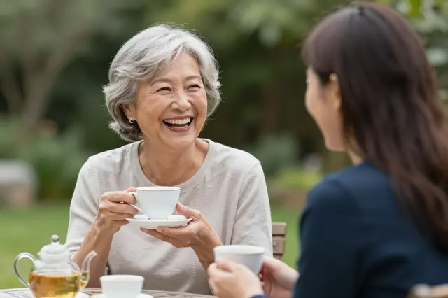 Laughing senior woman holds a teacup while sharing tea with a companion in a sunlit garden.