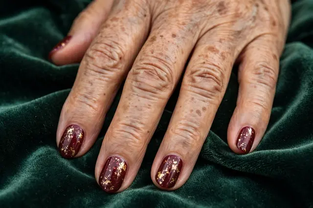 Close-up of an older woman's hand with burgundy gold-flake nail art resting on dark green velvet