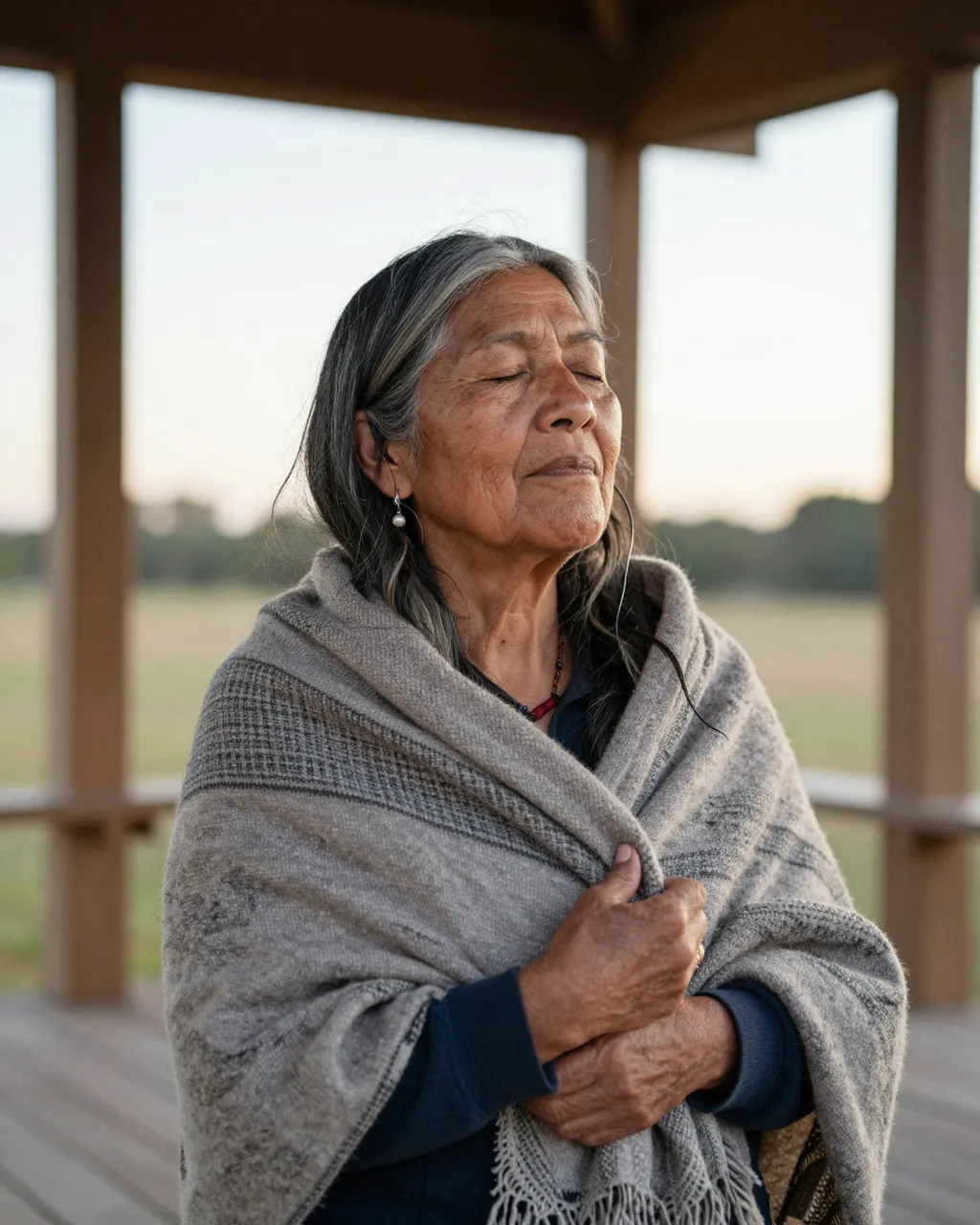 An older Indigenous woman with closed eyes wrapped in a woven shawl rests on a wooden porch at dawn.
