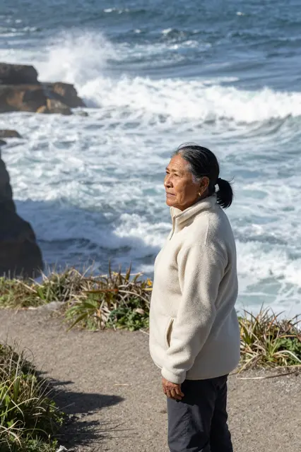 Side view of an older Indigenous woman in a cream fleece standing on a coastal path above crashing ocean waves