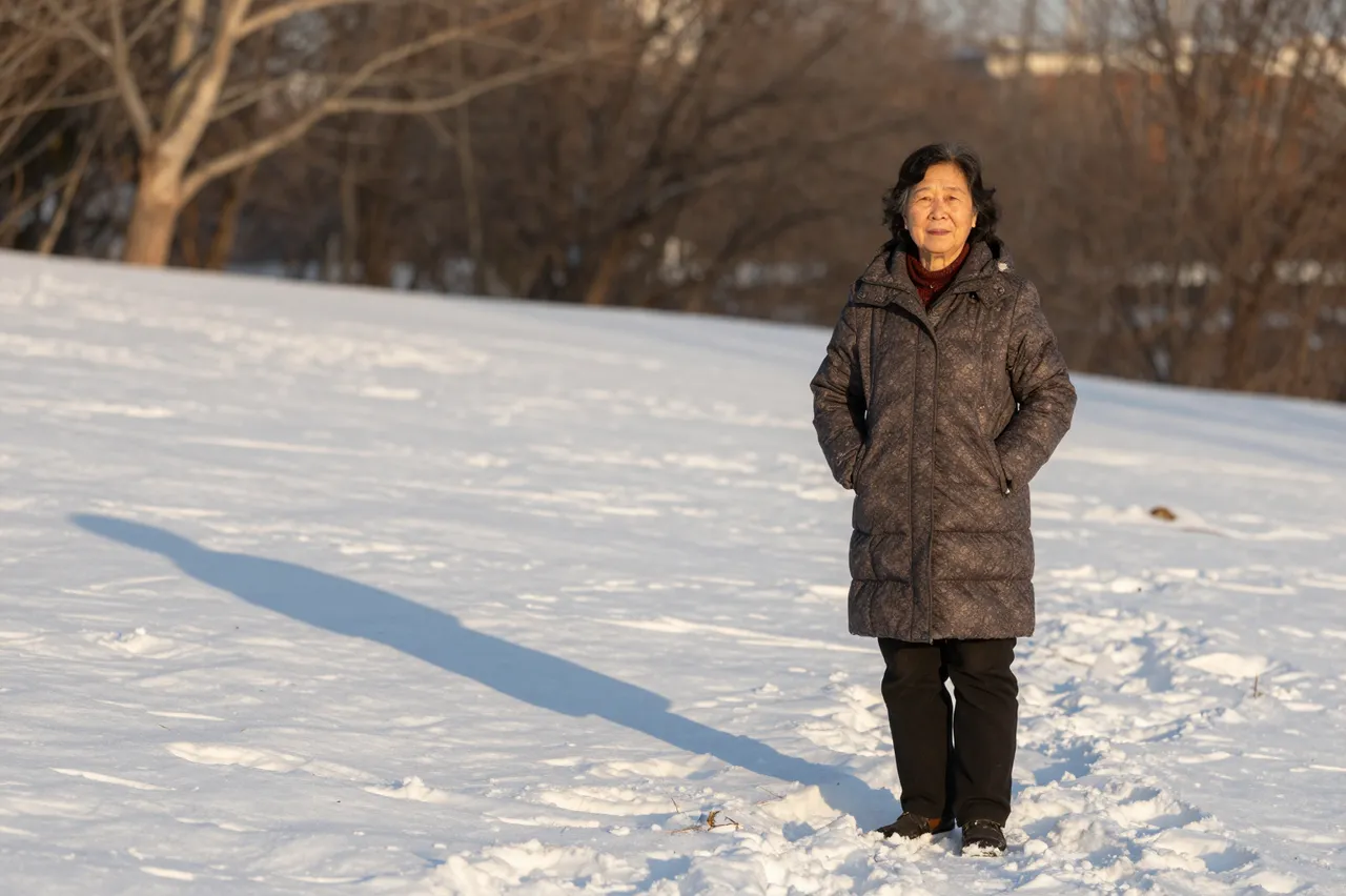 East Asian elderly woman in a winter parka stands on snow covered ground with a long golden hour shadow stretching beside her