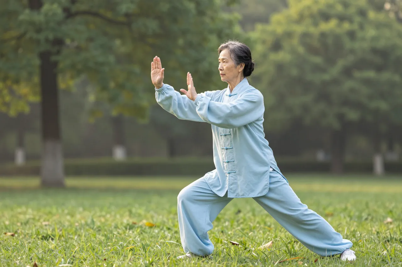 Elderly woman in light blue uniform performs tai chi on a grassy park lawn at dawn