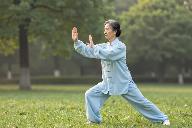 Elderly woman in light blue uniform performs tai chi on a grassy park lawn at dawn