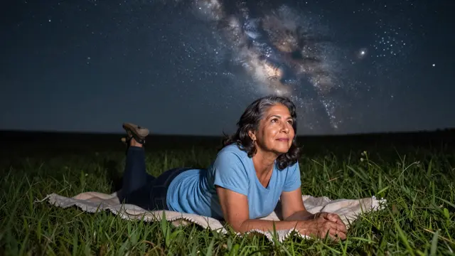 A Latina woman in her sixties lies on a blanket in a grassy meadow looking up at the Milky Way night sky.