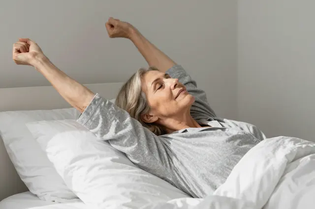 A woman in her sixties stretches her arms upward while waking up in a sunlit bedroom bed.